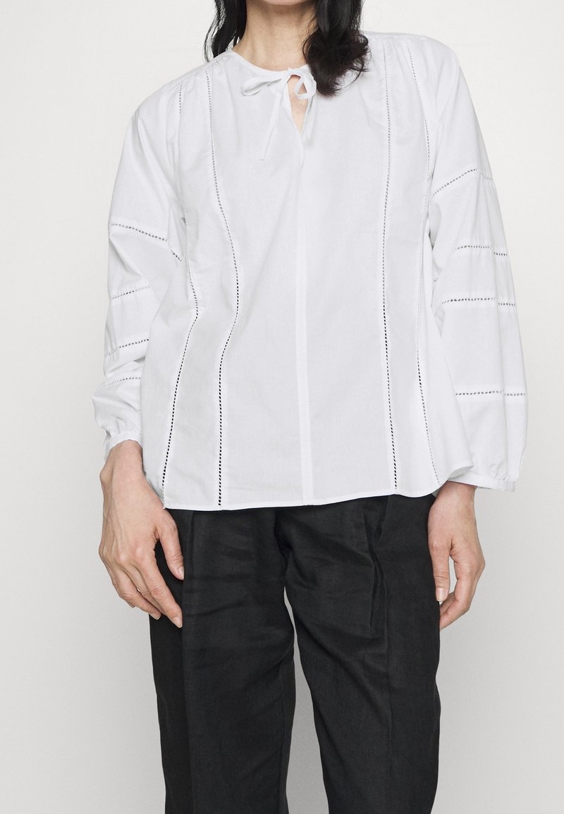Blusa bianca in cotone, con chiusura a fiocco, maniche lunghe a sbuffo, dettagli di cuciture verticali e un design a vestibilità dritta.
