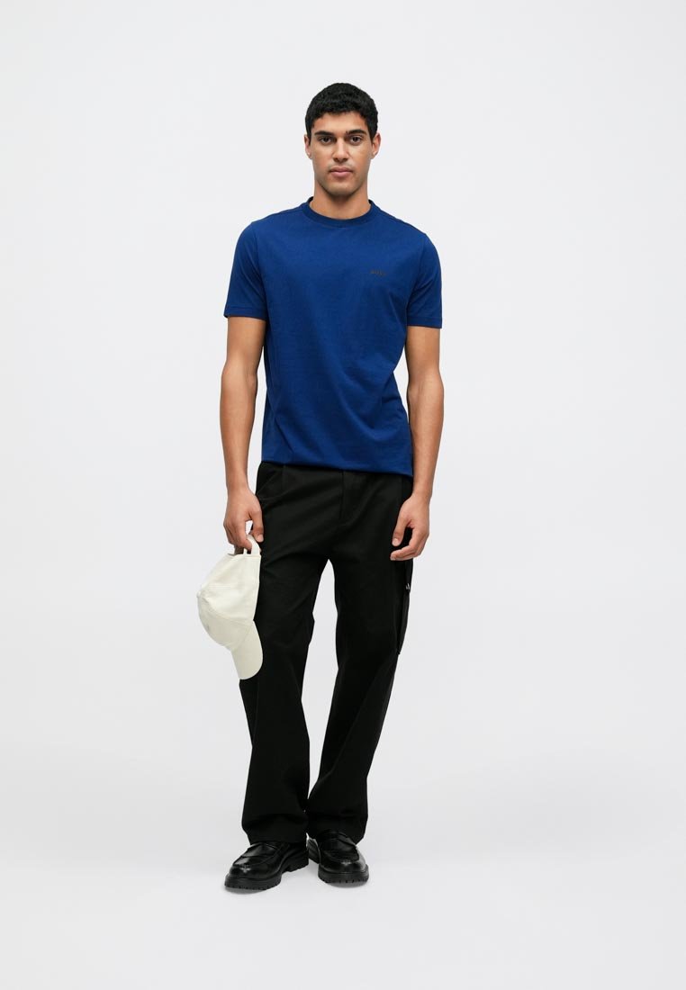 T-shirt bleu à manches courtes avec un col rond, associé à un pantalon noir et tenant une casquette de baseball blanche. Tissu lisse avec des détails minimaux.