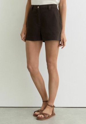 Frau, die dunkelbraune Shorts mit hoher Taille und braune Sandalen mit Knöchelriemen trägt und vor einer schlichten Wand steht.