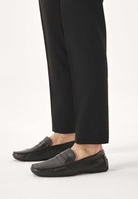 Mocasines de cuero negro con superficie texturizada y detalle de doble correa, combinados con pantalones negros. Diseño de perfil bajo con suelas de goma.
