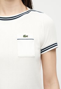 Haut en maille blanc à manches courtes avec une bordure rayée bleu marine au col et aux manches, avec une petite poche poitrine ornée d'un logo crocodile vert.