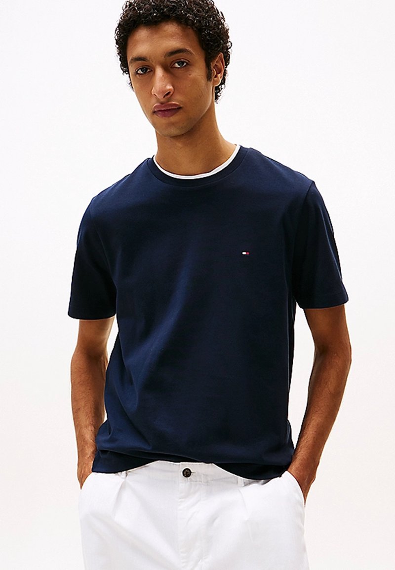 T-shirt di cotone blu navy con scollo rotondo classico, maniche corte e un piccolo logo sul petto, abbinato a pantaloni bianchi.