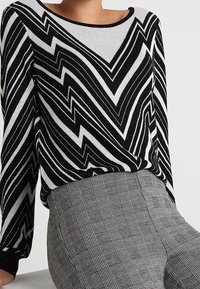 Schwarzer und weißer Pullover im Zickzackmuster mit rundem Ausschnitt, kombiniert mit grauen Karohosen. Der Pullover hat lange Ärmel und eine lockere Passform.