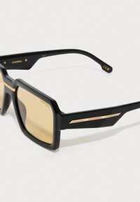 Óculos de sol retangulares pretos com detalhes em dourado. Armação espessa e mate com lentes castanhas claras. Apresenta uma marcação discreta nas hastes.