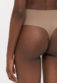 Close-up of a person’s lower back and hips wearing seamless beige thong underwear, showing natural skin texture and stretch marks.