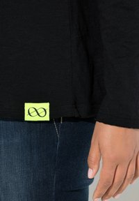 Schwarzes Langarmshirt mit einem auffälligen neongrünen Etikett, das ein Unendlichkeitssymbol zeigt, kombiniert mit dunkelblauen Jeans. Glatte Textur.