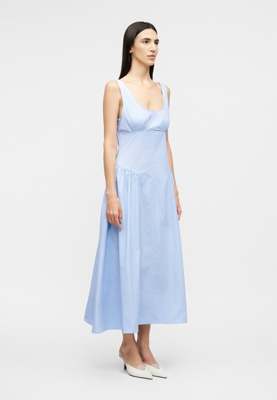 Γυναίκα που στέκεται, φορώντας ένα αμάνικο γαλάζιο φόρεμα μήκους μέχρι τη μέση της γάμπας με στρογγυλή λαιμόκοψη και λευκές μυτερές γόβες, μπροστά σε ένα απλό φωτεινό φόντο.