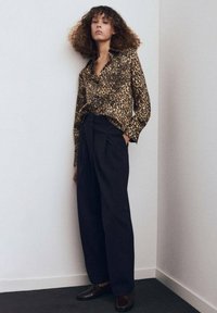 Camisa de estampado de leopardo hecha de tela ligera, metida dentro de pantalones negros de tiro alto con pliegues, combinada con mocasines oscuros.