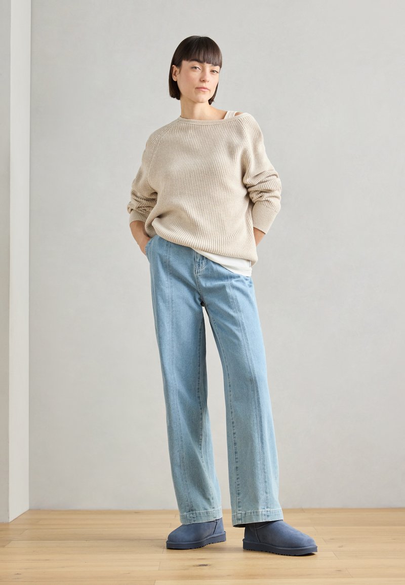 Beige gestrickter Pullover mit lockerer Passform, hellblaue gerade Jeans und blaue Stiefeletten, vor einem neutralen Hintergrund.