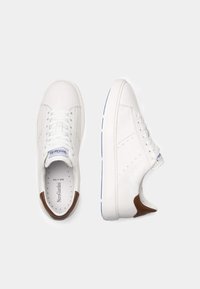 Weiße Lederturnschuhe mit glatter Oberfläche, braunen Akzenten an der Ferse, perforierten Seiten und flachen weißen Sohlen mit blauen Details.