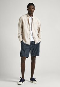 Camicia a maniche lunghe a quadri beige chiaro, t-shirt bianca con grafica, pantaloni corti blu scuri, sneakers blu con suola bianca, in piedi contro uno sfondo neutro.