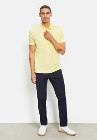 Geel polo shirt met korte mouwen gemaakt van gestructureerde stof, gecombineerd met smal gesneden marineblauwe broek en witte sneakers. Model staat met de hand op de kraag.