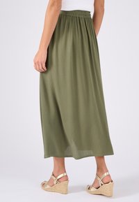 Falda verde oliva hasta los tobillos con una cintura fruncida y textura vertical. Combinada con sandalias de cuña doradas con un diseño de tiras.