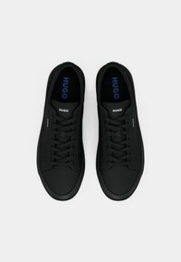 Μαύρα αθλητικά παπούτσια με λεία, ματ φινίρισμα, που διαθέτουν στρογγυλά κορδόνια, στρογγυλή μύτη και το όνομα της μάρκας "HUGO" σε μπλε στα εσωτερικά τους.