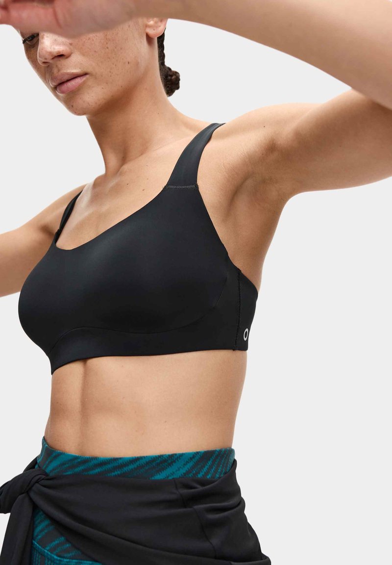 Zwarte sportbeha van glad materiaal, met brede bandjes en een naadloos ontwerp. Heeft minimale stiknaden en een klein logo nabij de zijkant.