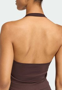 Hnědý top na zavazování za krk s přiléhavým střihem, žebrovanou texturou a otevřenými zády, které odhalují hladkou pokožku na ramenou a krku.