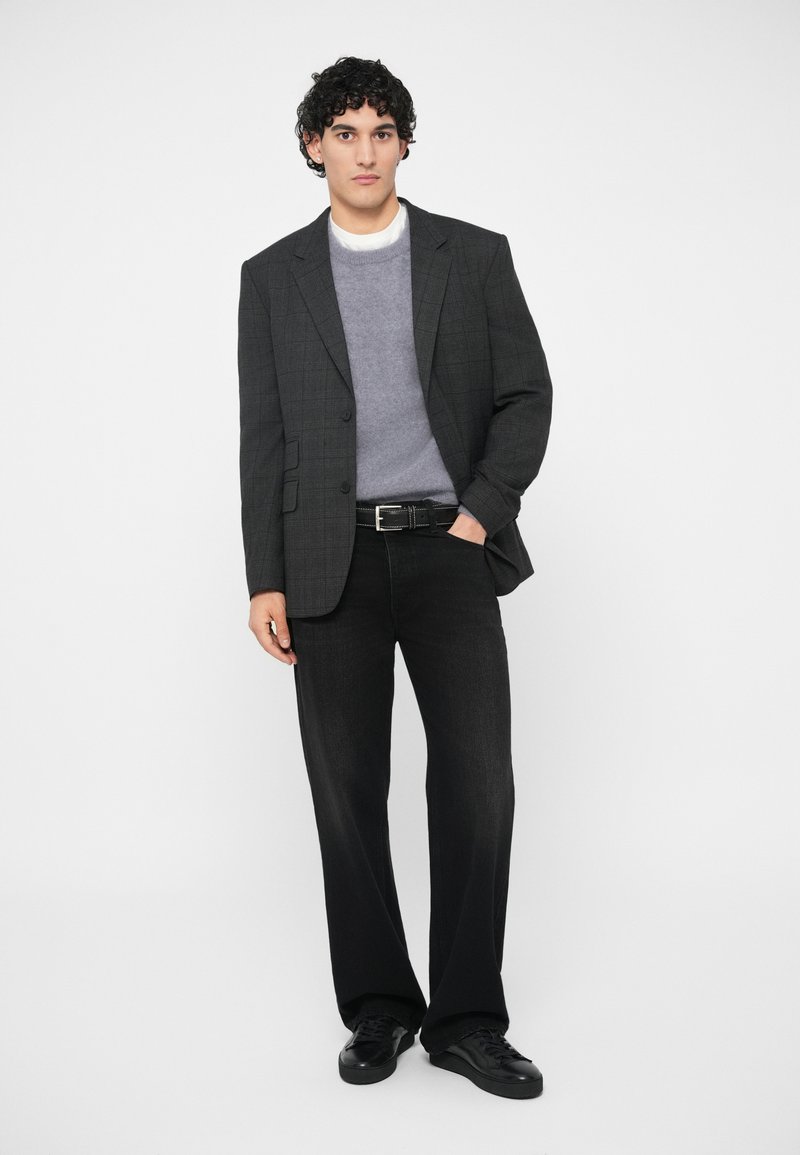 Mann in einem dunklen karierten Blazer, grauem Pullover über einem weißen Hemd, schwarzem Gürtel, schwarzen weit geschnittenen Jeans und schwarzen Sneakers, der mit einer Hand in der Tasche steht.