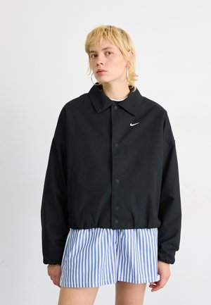 Schwarze Nike-Jacke mit Kragen und Druckknopfverschluss, ausgestattet mit Seitentaschen und dezentem Logo; kombiniert mit blau-weiß gestreiften Shorts.