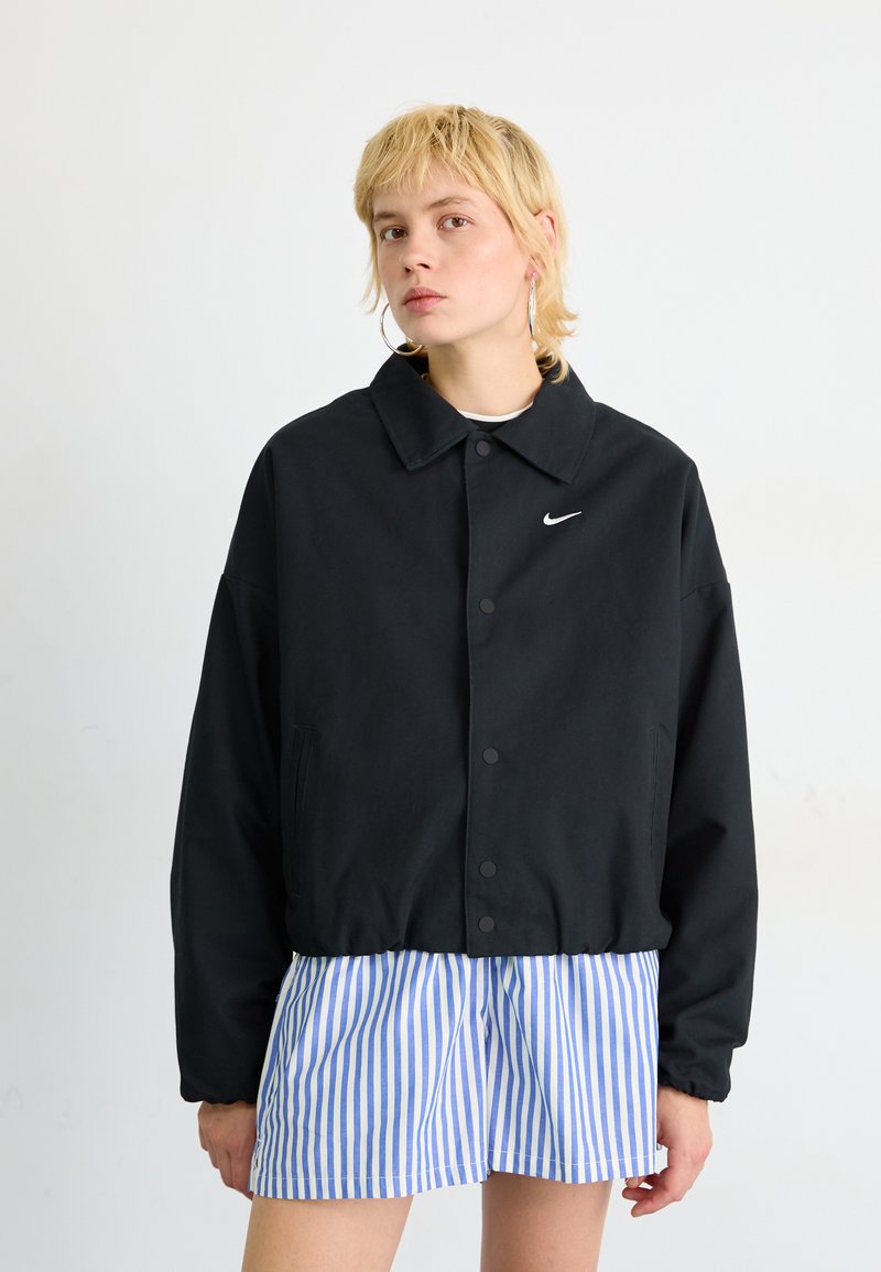 Crna Nike jakna s kragnom i kopčanjem na dugmad, s bočnim džepovima i suptilnim logotipom; uparena s plavim i bijelim prugastim kratkim hlačama.