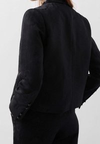 Musta teksturoitu bleiseri räätälöidyllä istuvuudella, jossa on takasauma, napitettu hihansuu ja hienovaraisia kuvioita. Sujuva kankaan pinta.