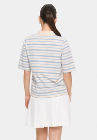 Polo à manches courtes en crème avec des rayures horizontales bleues et beige. Il dispose d'un col et d'une coupe décontractée. Associé avec une jupe plissée blanche.