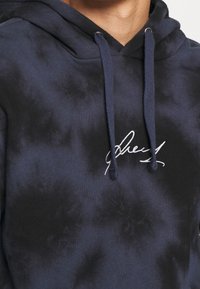 Felpa navy tie-dye con cappuccio regolabile e logo bianco ricamato. Tessuto morbido con una consistenza liscia. Presenta cuciture rinforzate.