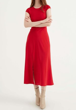 Robe rouge jusqu'au genou avec un corsage ajusté, des manches courtes et une fente à l'avant. Tissu lisse avec un léger éclat ; portée avec des bottines beiges.