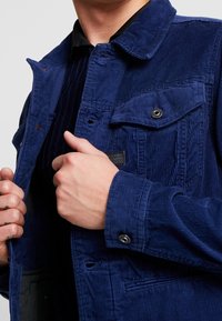 Blå kordjacka med krage, framfickor och tryckknappar. Den texturerade tyget har vertikala ribbor och en lätt glänsande yta.