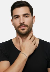 Bracciale a catena d'argento su una mano, indossato con una maglia nera a scollo a V. Primo piano dei tratti del viso, rasato e con capelli corti e scuri.