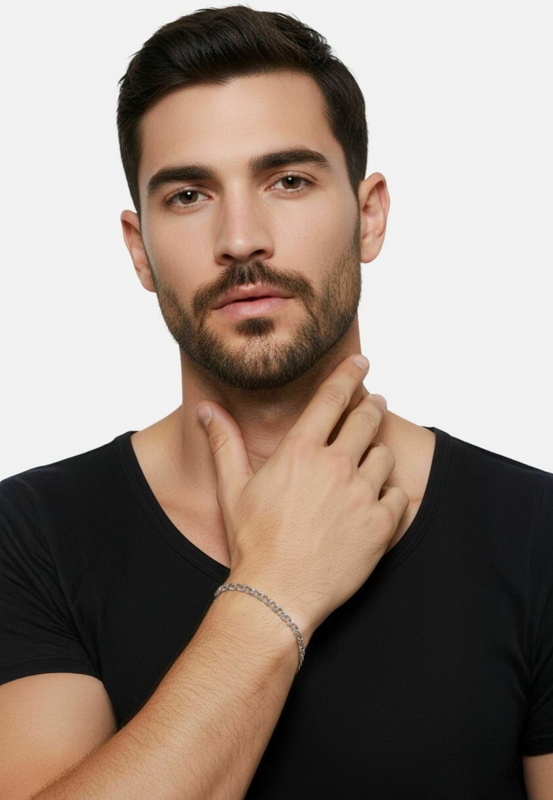 Bracciale a catena d'argento su una mano, indossato con una maglia nera a scollo a V. Primo piano dei tratti del viso, rasato e con capelli corti e scuri.