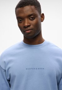 Sudadera azul claro hecha de tela suave, con cuello redondo y el logotipo "SCOTCH & SODA" impreso en azul más oscuro en el pecho.