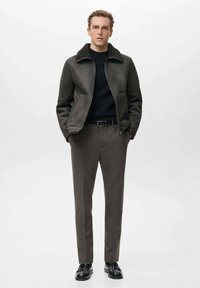 Giacca da uomo in shearling grigio scuro con colletto alto, abbinata a pantaloni grigi slim-fit e scarpe in pelle nere. Design minimalista.
