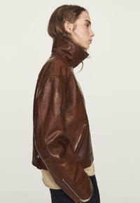 Braune Lederjacke mit hohem Kragen, seitlichen Reißverschlusstaschen und entspannter Passform. Die Struktur ist glatt mit einem dezenten Glanz.