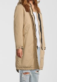Beige polstret parka-jakke med hætte, frontlynlås, sorte detaljer og sidelommer; har en afslappet pasform og en knælængde design.
