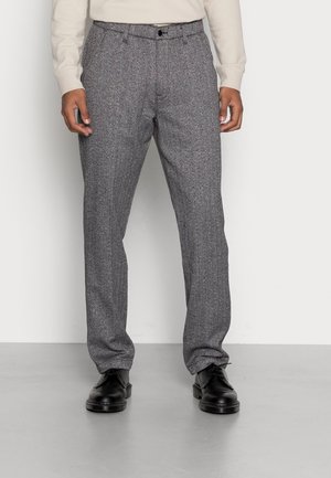 Mężczyzna ubrany w szare spodnie w jodełkę, czarne skórzane eleganckie buty i kremową koszulę z długim rękawem, stojący na jednolitym tle.