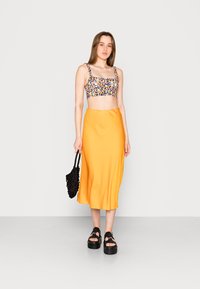 Top cropped floral com decote quadrado, combinado com uma saia midi laranja sólida. Modelo usa sandálias pretas de plataforma e segura uma bolsa de palha preta.