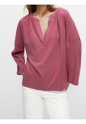 Vrouw draagt een losse roze blouse met lange mouwen en een gespleten halslijn, gecombineerd met een witte broek, staand tegen een effen achtergrond.