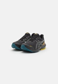 Pár černých běžeckých bot ASICS s modrými podrážkami, žlutými akcenty a brandingem Gore-Tex voděodolnosti na bílém pozadí.