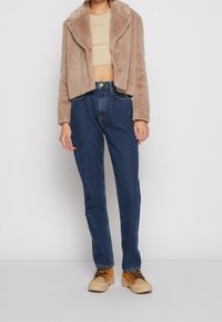 Person som bär en kort beige imitation av pälsjacka, beige kort topp, högmidjade blå jeans och beige snörstövlar, stående mot en vit bakgrund.