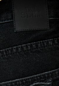 Černá denimo tkanina s úzkým střihem, obsahující texturovanou černou koženou nášivku s nápisem "THE DENIM." Viditelné švy podél švů.