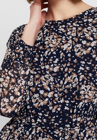 Floral dress featuring a navy background with tan and white patterns, long sleeves with button accents, and a fitted waist with a tie.