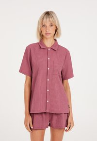 Camisa de manga corta con cuello en un profundo color rosa, de tela texturada, con un bolsillo en el pecho izquierdo y botones blancos. Combinada con pantalones cortos a juego.