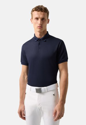 Ung mand iført en marineblå poloshirt og hvide bukser med et hvidt bælte, stående med fronten mod kameraet foran en ensfarvet baggrund.