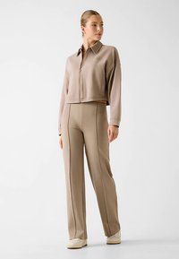 Beige gekürzte Reißverschlussjacke mit Kragen, kombiniert mit beigefarbenen, geraden Hosen mit Seitennähten. Glatte Textur, minimale Details.