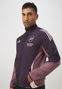 Lila Sportjacke mit hohem Kragen, Reißverschlussfront und kontrastierenden hellrosa Seiteneinsätzen. Logo auf der Brust und Taschen an den Seiten.