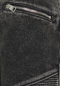 Detailní záběr na černou denimovou látku se stříbrným zipem, který je částečně zapnutý na malé kapse.