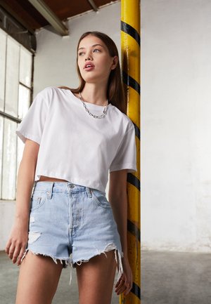 Witte cropped t-shirt, losse fit, gecombineerd met lichtblauwe versleten denim shorts met verweerde randen. Op de achtergrond staat een gestreepte paal.