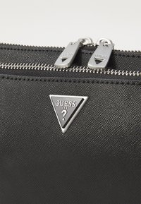 Borsa a mano in materiale sintetico nero e textures, con cerniere argentate e un'etichetta a logo metallico triangolare che presenta "Guess" e un punto interrogativo.