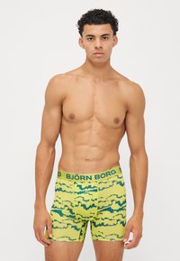 Κίτρινες μποξεράκιοι με μπλε αφηρημένα σχέδια, διαθέτουν ελαστική μέση με την επιγραφή "BJÖRN BORG." Μαλακός, εφαρμοστός σχεδιασμός με μήκος μέχρι το μέσο μηρού.