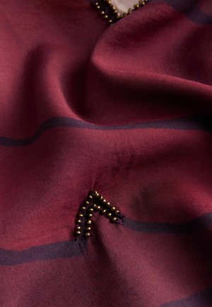Zbliżenie pomarszczonej ciemnoczerwonej tkaniny z czarnymi poziomymi paskami i małymi ozdobami z mosiężnych koralików tworzącymi częściowy trójkąt.